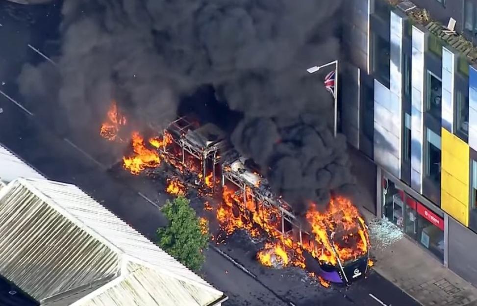 A double-decker bus engulfed in flames and black smoke on a street in Belfast.