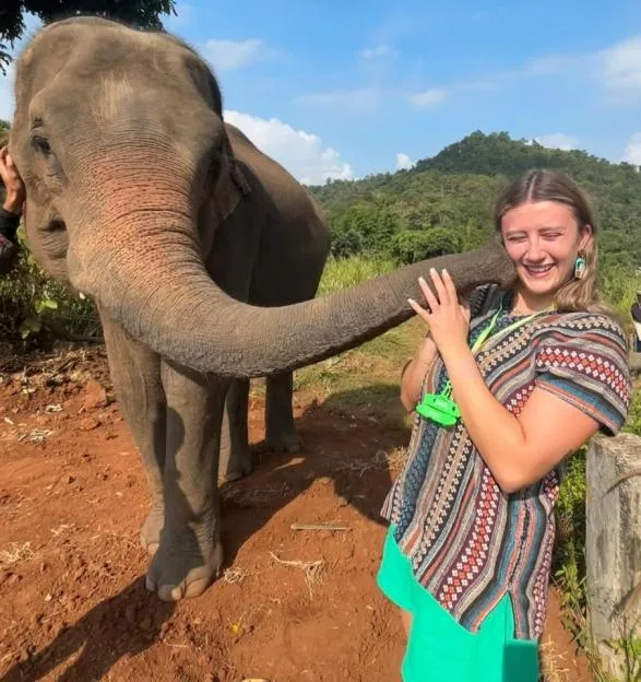 A smiling young woman standing outdoors, laughing as an elephant wraps its trunk playfully around her neck.