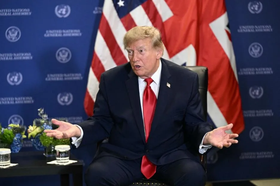 Donald Trump speaks with hands open, wearing a dark suit and red tie, in front of American and British flags and a UN backdrop.