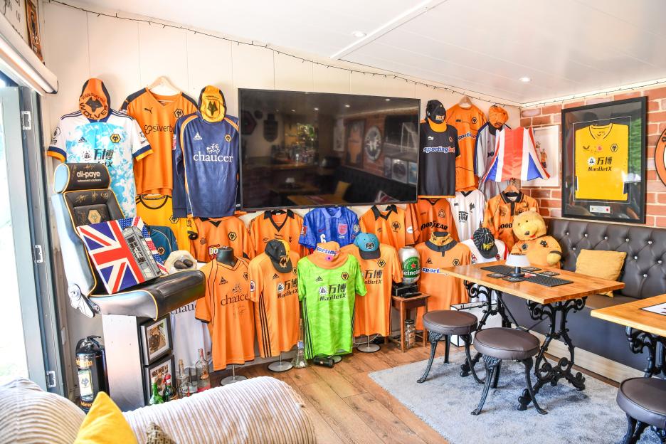 A sports bar in a backyard shed with Wolverhampton Wanderers FC memorabilia, including many jerseys, a large TV, pub benches, and a dartboard.