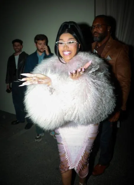 Cardi B in a light pink furry top and a light pink skirt, with two men behind her.
