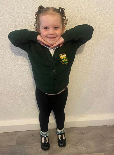 A young girl in a school uniform, green cardigan with a school crest, and black leggings, smiles while posing with her hands under her chin.