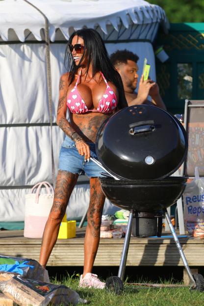 Katie Price, in a pink heart-print bikini top and denim shorts, stands by a barbecue with a man in the background.