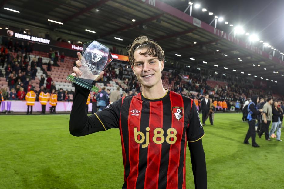 A male soccer player in a red and black striped jersey smiles while holding up a "Supporters' Player of the Season" award at a stadium.