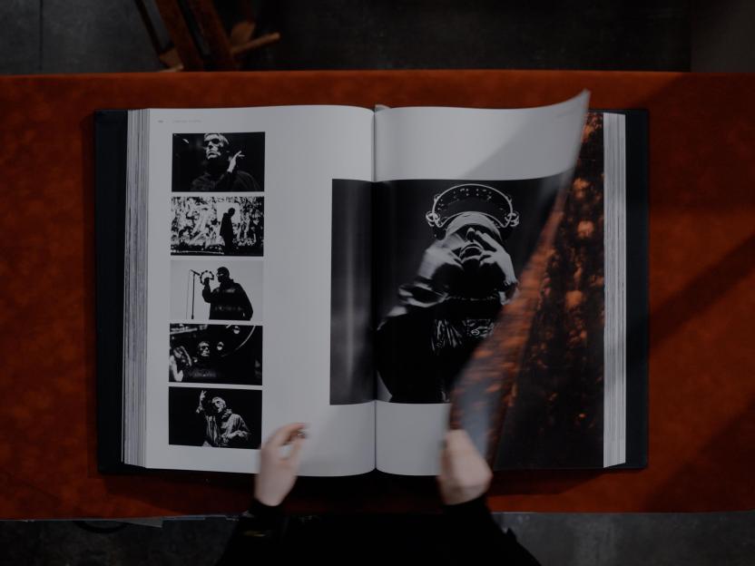 Hands flipping through "The Oasis Live '25 Opus" book, showing black and white photos of the band performing.