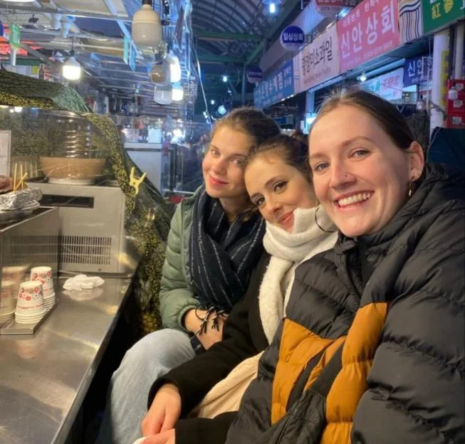 Three young women smiling together at a food stall in a bustling night market.