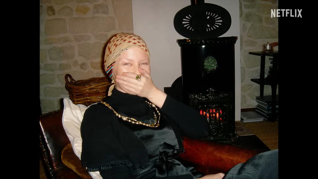 Kylie Minogue sitting on a couch with a headscarf on, holding her hand over her mouth.