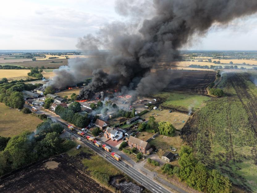 Aerial view of a large fire consuming buildings, with massive plumes of black smoke rising into the sky and fire trucks on a nearby road.
