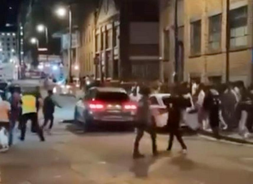 Video still of a black car speeding through a crowd of people on a city street at night.