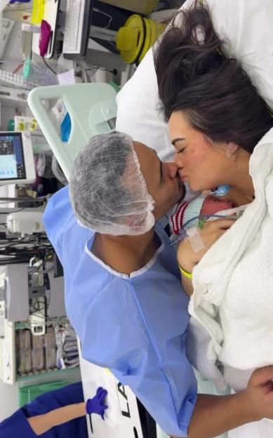 A new mother lying on a hospital bed and a man in a surgical gown and cap are kissing with a newborn baby between them.