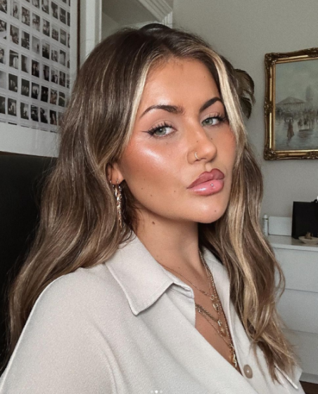 A young woman with highlights in her long hair, wearing a white button-up shirt and layered necklaces, looks at the camera.