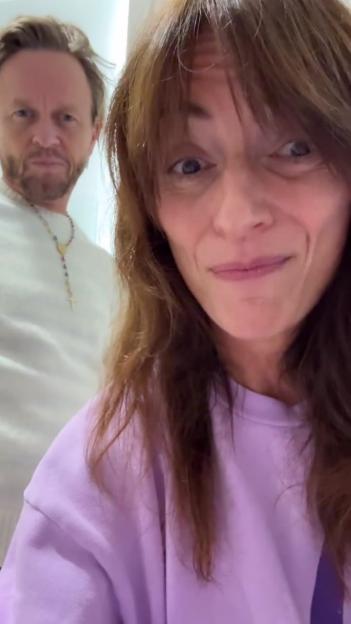 A woman in a purple top in the foreground smiling while a man in a white top with a rosary necklace looks on in the background.