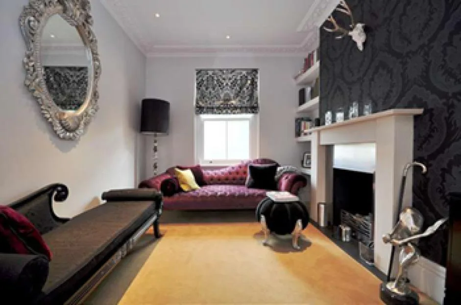 An interior view of a room with a fireplace, a large ornate mirror, a magenta velvet daybed, and a yellow rug.