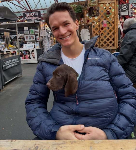 A smiling man holding a brown puppy inside his jacket.
