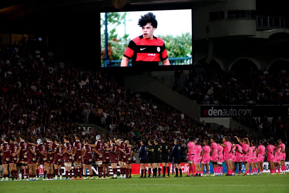 Rugby players from Stade Français and Bordeaux stand on the field facing a large screen displaying a player.