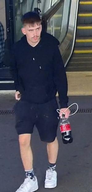 A man with a mustache, wearing a black hoodie and shorts, holds a partially filled Coca-Cola bottle.