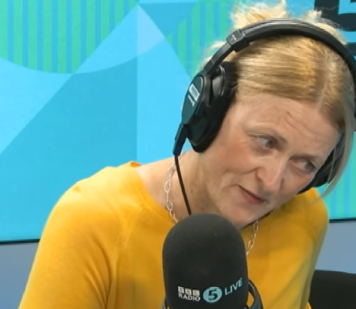 A woman with blonde hair, wearing headphones and a yellow shirt, looks to her right while speaking into a microphone.