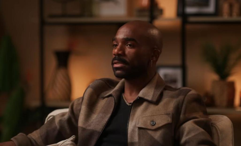 Ore Oduba, a balding man with a mustache and beard, wearing a plaid jacket and a gold chain.