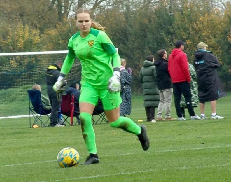 A female soccer player in a green uniform and muddy knees runs after a soccer ball.