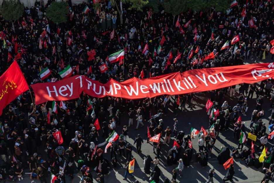 An overhead shot of a large crowd of mourners dressed in black carrying flags and a red banner that reads "Hey Trump We will kill You".