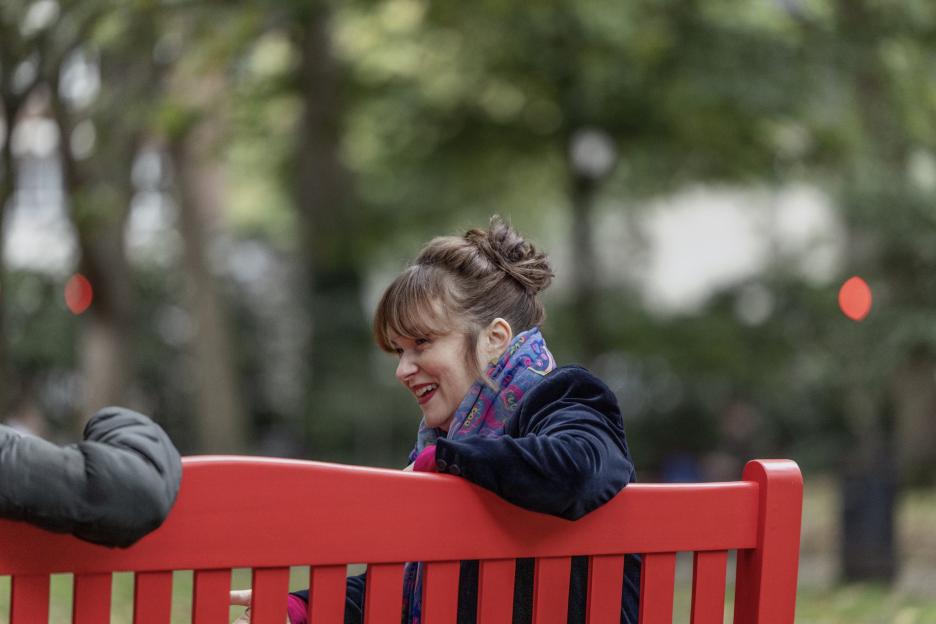 A woman with her hair in a bun and a colorful scarf around her neck sitting on a red park bench and smiling.