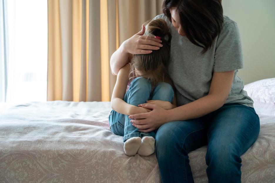 A mother consoles her crying daughter who is sitting on a bed.