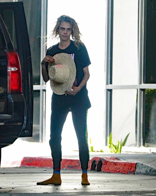 Cara Delevingne in a black shirt and black pants, holding a straw hat, standing barefoot outside a private jet.