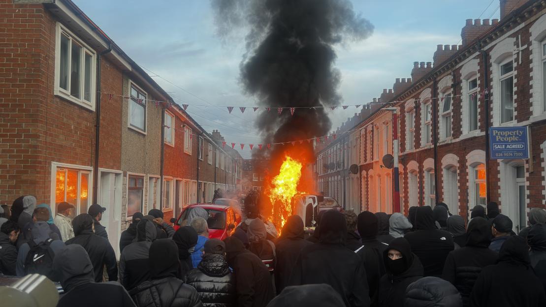 Protesters gathered on Lendrick Street in Belfast as a vehicle burns in the street, sending large amounts of black smoke into the sky.