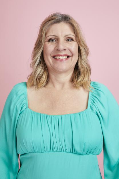 Kari Roberts smiling, wearing a teal dress with a square neckline, against a pink background.