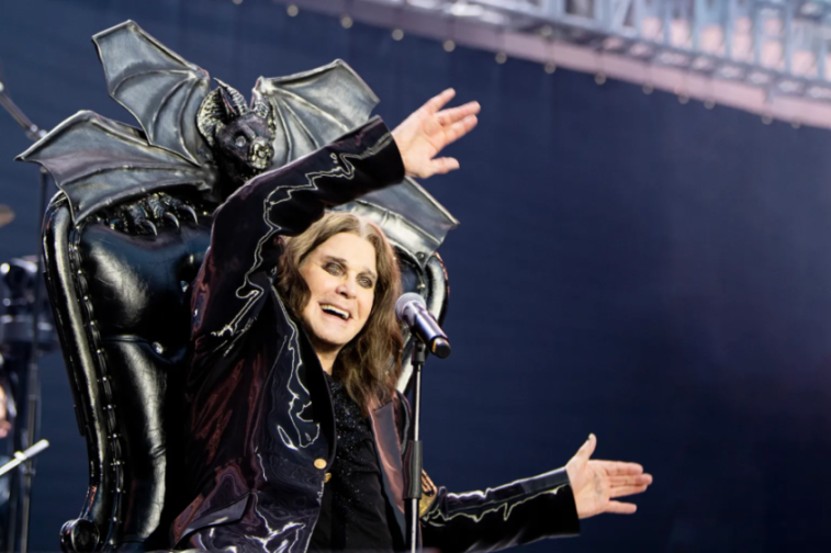 Ozzy Osbourne in a black suit, sitting in a black chair with a bat-like figure on top, singing into a microphone with his arms outstretched.