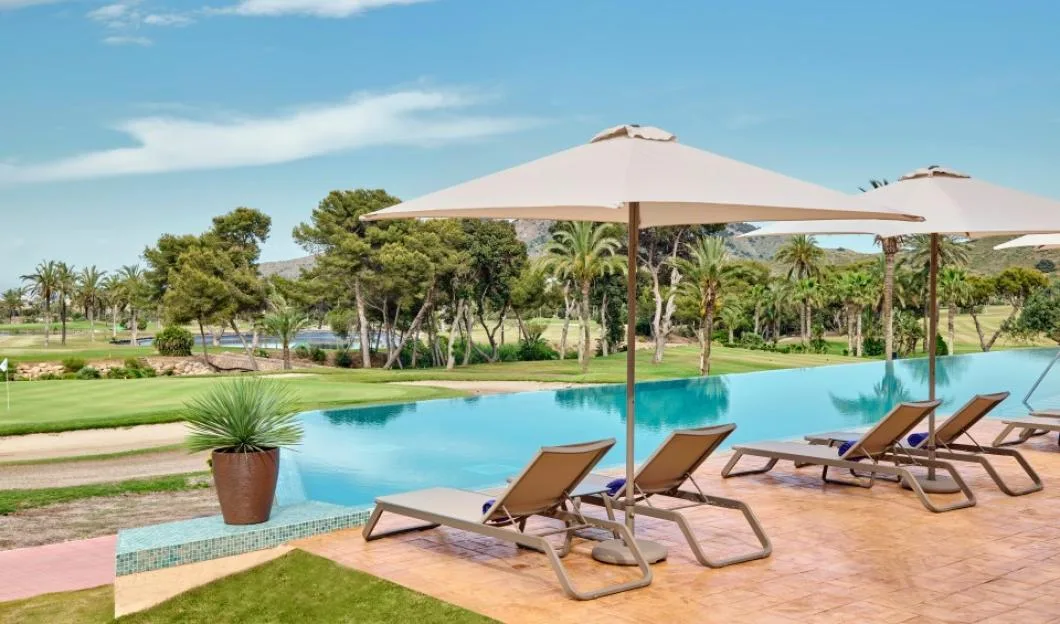 Sun loungers and umbrellas beside an infinity pool overlooking a golf course and palm trees.