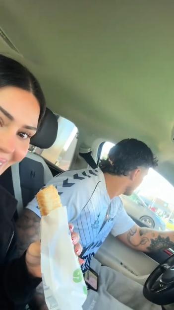 A woman holds a pastry in a car while a man with a mullet and tattoos sits in the driver's seat.