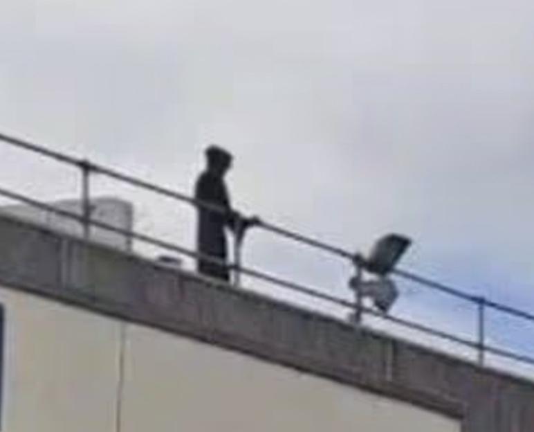 A person in a dark hooded garment stands on a rooftop with a railing.
