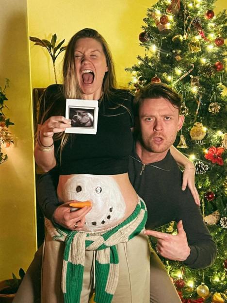 A couple stands in front of a Christmas tree, with the woman displaying an ultrasound picture and her belly painted like a snowman, while the man points at it.
