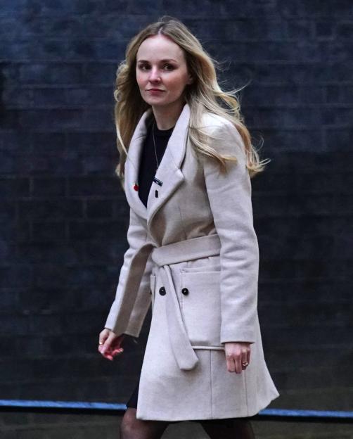 Lissie Harper walking, wearing a light-colored coat with a poppy pin.
