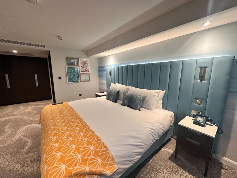 A bedroom in the new Hotel Indigo Torquay by IHG with a blue upholstered headboard, white bed with orange throw, and framed art on the wall.