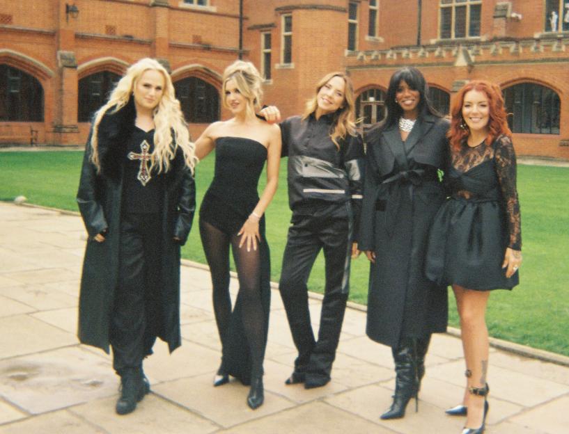 Rebel Wilson, Ashley Roberts, Mel C, Shaznay Lewis, and Sheridan Smith posing together outdoors in front of a brick building.