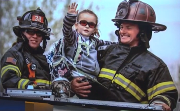 A young girl in sunglasses sits between two firefighters on a fire truck, waving.