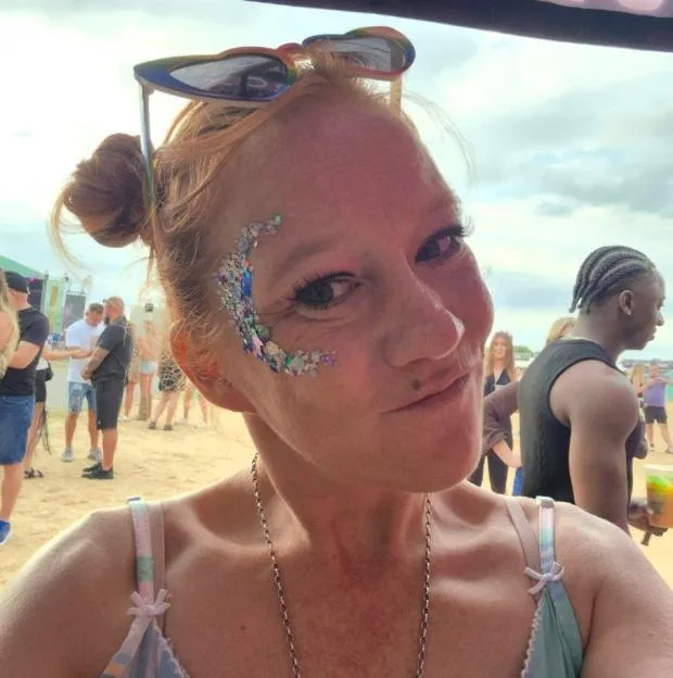 Amanda Martin smiles for a selfie at an outdoor event, wearing glitter face makeup, sunglasses on her head, and a patterned top.