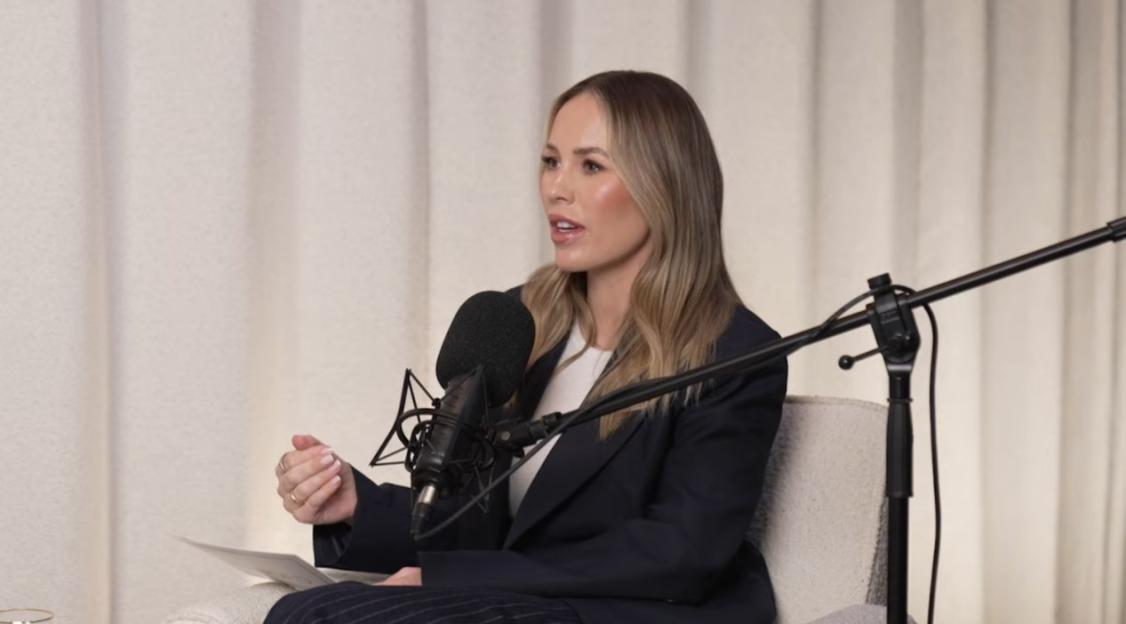 A woman with long blonde hair and wearing a dark blazer speaking into a microphone.