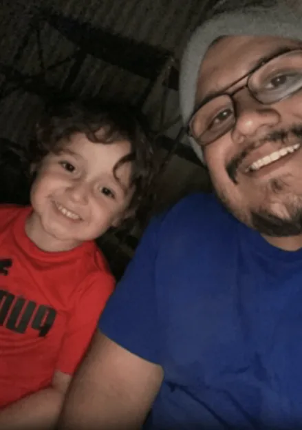 A young boy with curly hair in a red shirt smiles next to a man in a blue shirt, glasses, and a gray beanie.