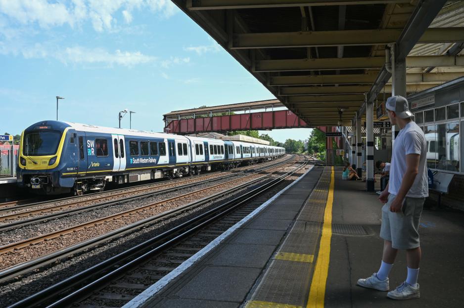 A man in a white t-shirt and grey shorts waits on a train platform with a train stopped at the adjacent tracks.