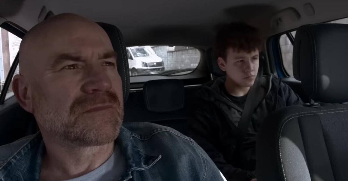 A bald man with a beard driving a car while a young man sits in the back seat.