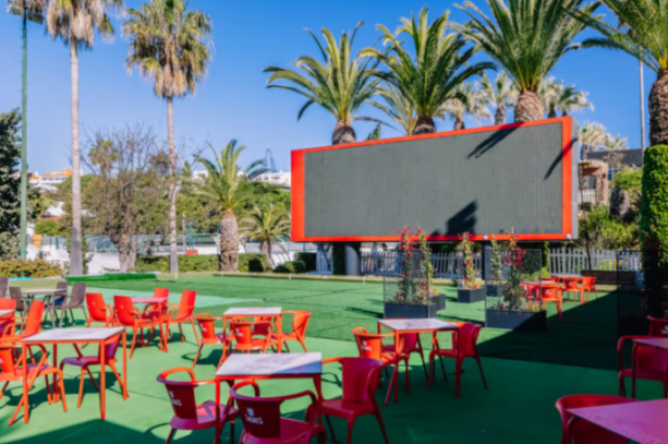 Outdoor dining area at Muthu Clube Praia da Oura with red tables and chairs, palm trees, and a large empty screen.