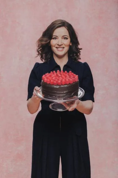 A smiling woman in a dark blue top and pants holds a chocolate cake topped with raspberries on a clear cake stand.