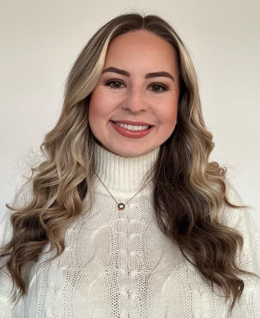 A smiling woman with long, wavy blonde and brown hair wearing a white turtleneck sweater and a delicate gold necklace.
