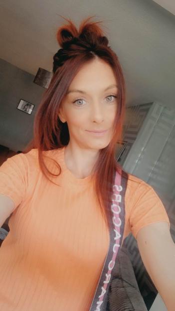 Vicky Waugh, a woman with auburn hair in a messy bun, smiles while wearing an orange t-shirt and a shoulder bag.