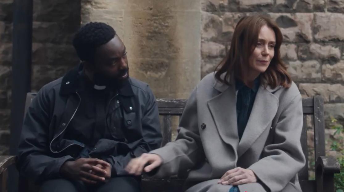 A Black man in a clerical collar and a white woman with brown hair sit on a bench, looking at each other.