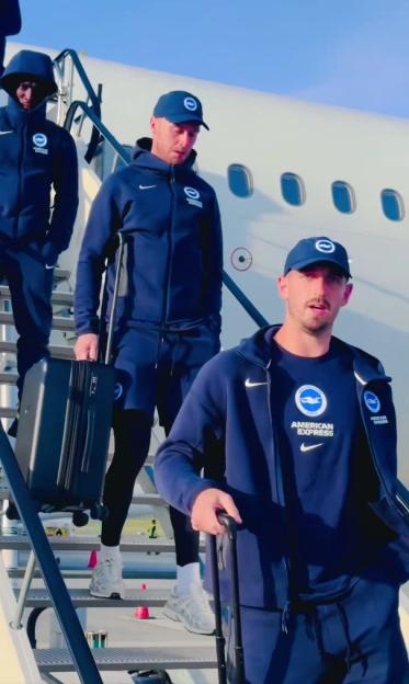 Three men in navy tracksuits with "Brighton & Hove Albion" and "American Express" logos descending airplane stairs with luggage.