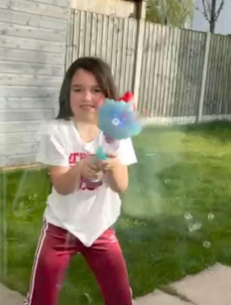A young girl in a white t-shirt and red pants smiles while holding a bubble gun that is blowing bubbles in a grassy backyard.